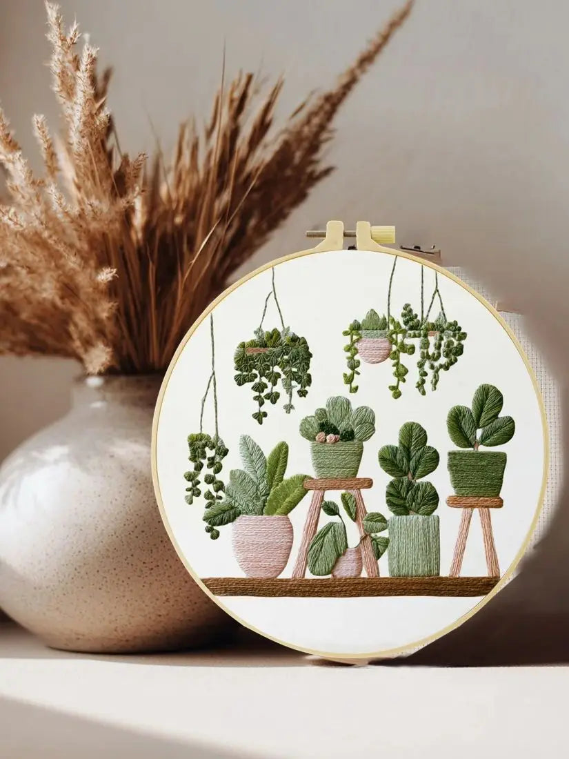 A cross stitch kit featuring green potted plants hanging from strings in a circular embroidery design.