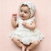 A baby girl wearing a white lace tulle dress and matching hat, sitting on a pink background.