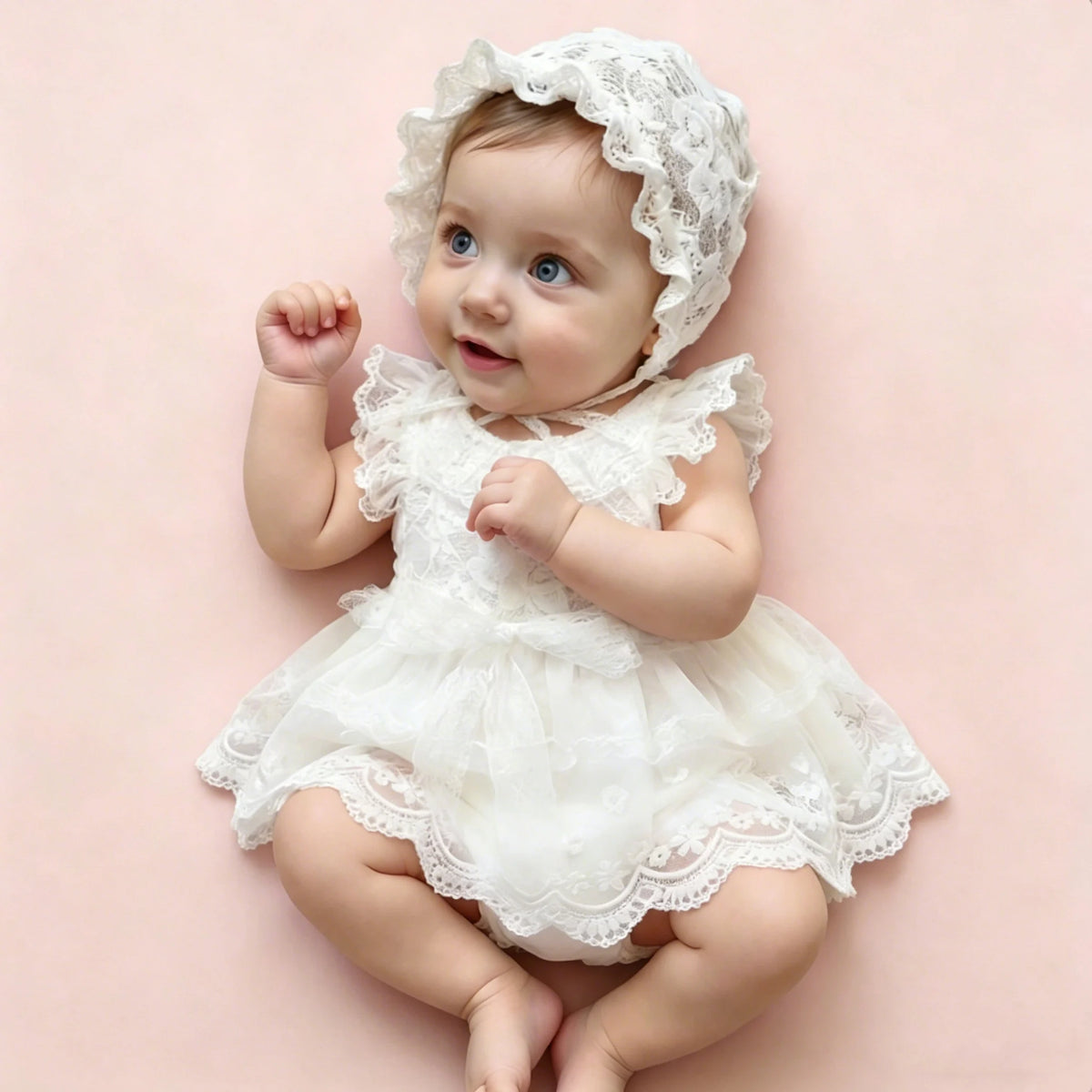 A baby girl wearing a white lace tulle dress and matching hat, sitting on a pink background.