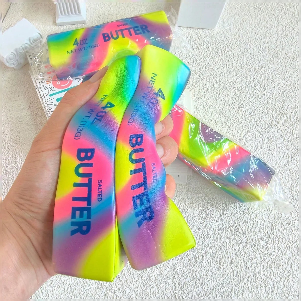 A hand holds a neon butter stick squishy toy in various colors, demonstrating its soft texture and vibrant hues.