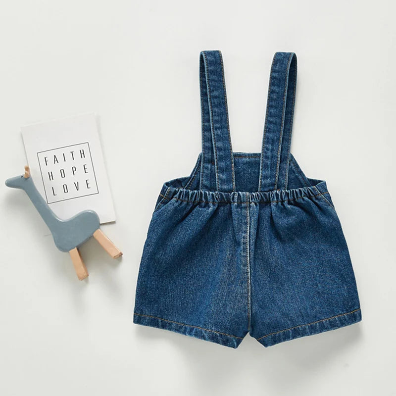 A pair of dark blue denim toddler romper shorts with vertical straps and pockets.