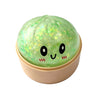 A translucent green squishy fidget toy with glitter and simple black eyes resting in a light beige container.