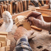 A hand carves a wooden block using a knife in a workshop setting with other wood blocks visible.
