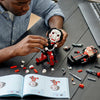 A person is assembling a black and white horror movie puppet model kit on a dark table next to a laptop and various small par