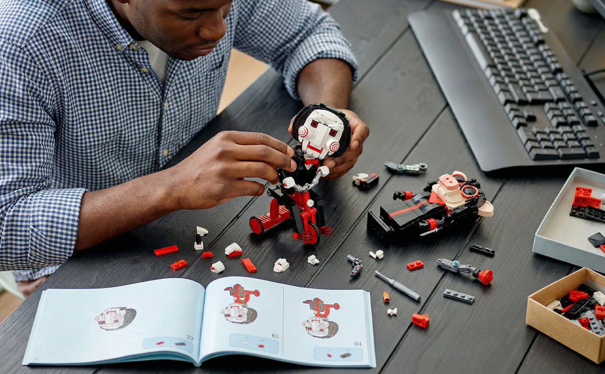 A person is assembling a black and white horror movie puppet model kit on a dark table next to a laptop and various small par