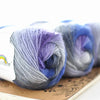A collection of hand-dyed merino wool yarn in shades of blue, purple, and gray rolled on a wooden surface.