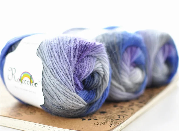 A collection of hand-dyed merino wool yarn in shades of blue, purple, and gray rolled on a wooden surface.