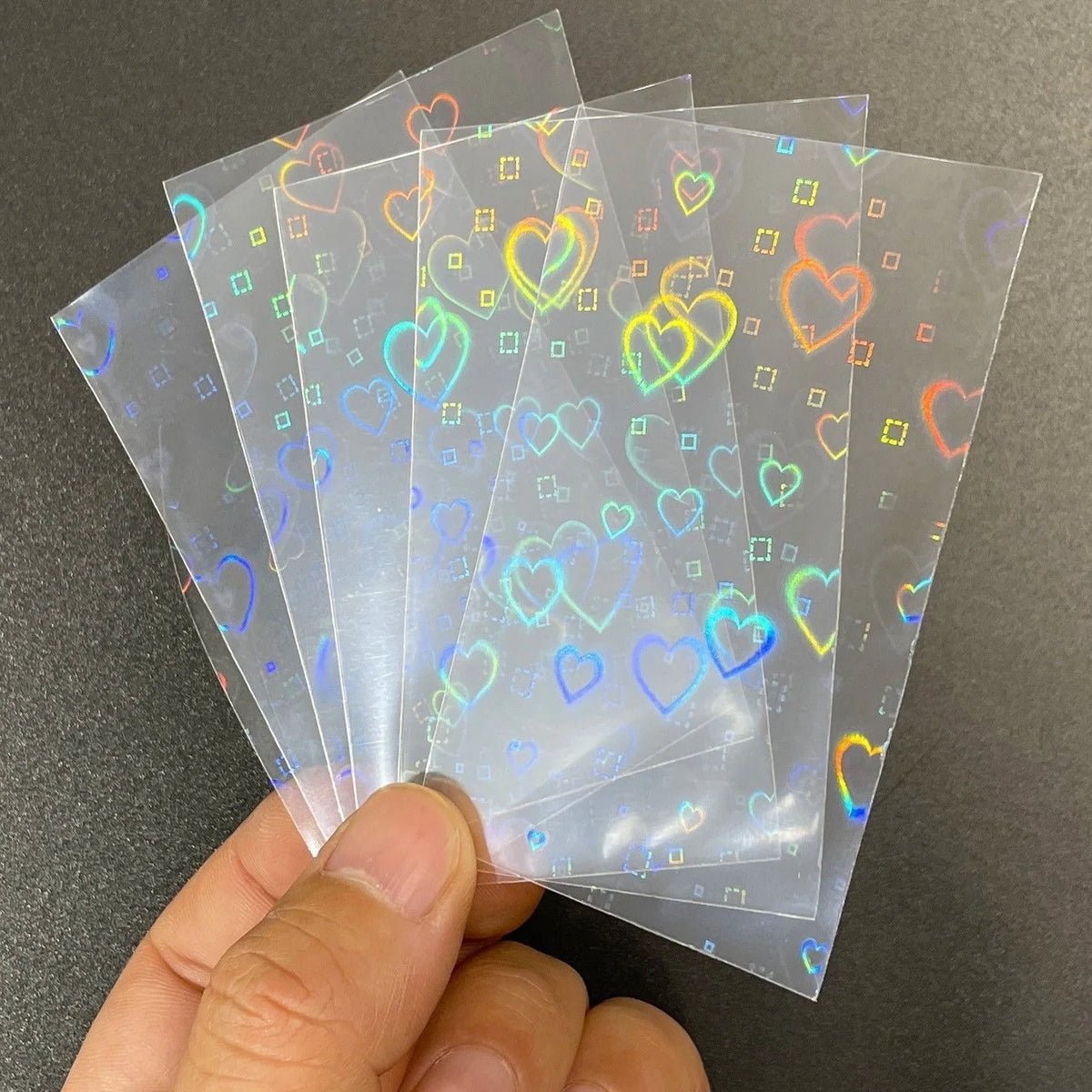 A hand holds a stack of holographic card sleeves for trading card games, showing various colors and heart designs.