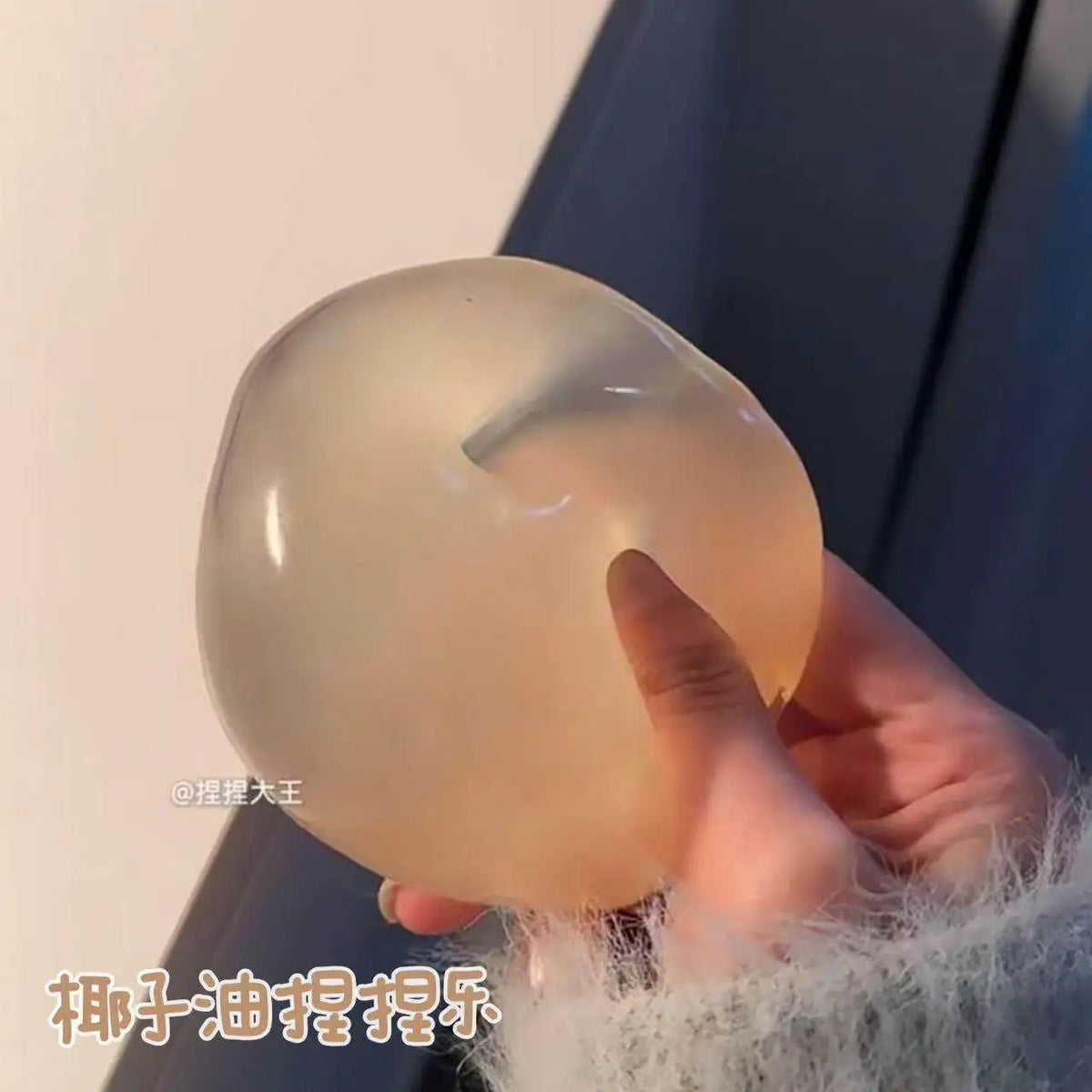 A hand is holding a smooth, translucent, light beige squeeze ball made of coconut oil for stress relief.