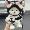 A white and black cow-print plush bear with pink ears and a black hood being held by a hand.