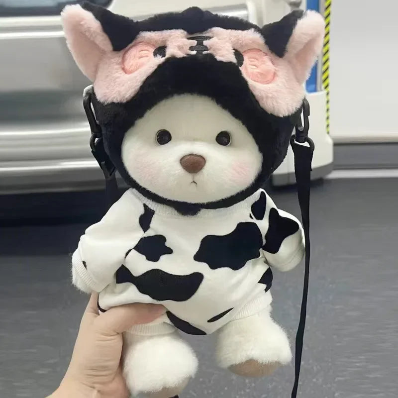 A white and black cow-print plush bear with pink ears and a black hood being held by a hand.