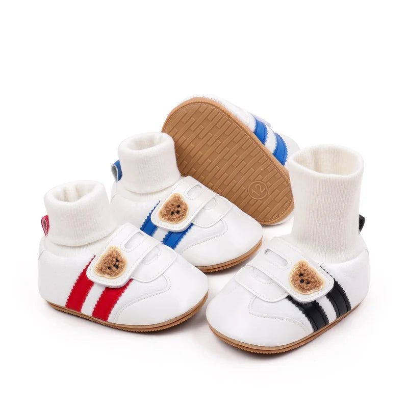 A pair of white ankle-hugging baby booties with anti-slip soles featuring red and blue stripes and brown cork.