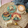 A set of eight DIY diamond art coasters and a holder are displayed on a woven mat with a cup of latte nearby.