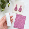 A pair of pink clay texture rollers and a finished piece of jewelry are displayed on a white surface.