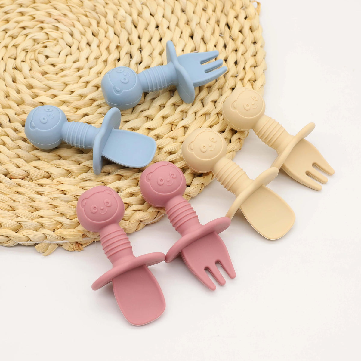 A set of nine silicone baby utensils including forks and spoons in various pastel colors resting on a woven mat.
