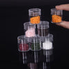 A hand holds several clear glass storage bottles filled with various colored seeds and grains like orange, pink, brown, green