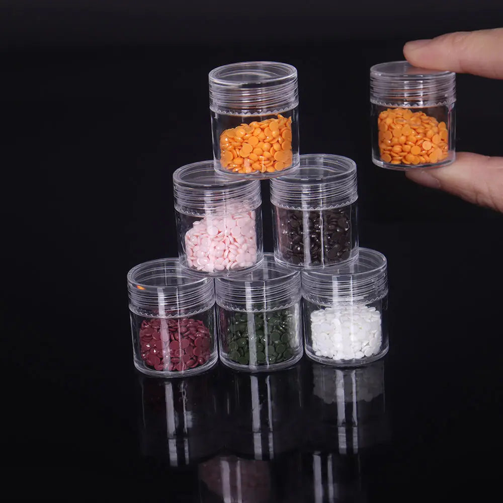 A hand holds several clear glass storage bottles filled with various colored seeds and grains like orange, pink, brown, green
