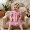 A toddler girl wearing a pink gingham A-line dress with a Peter Pan collar sitting on a sofa.