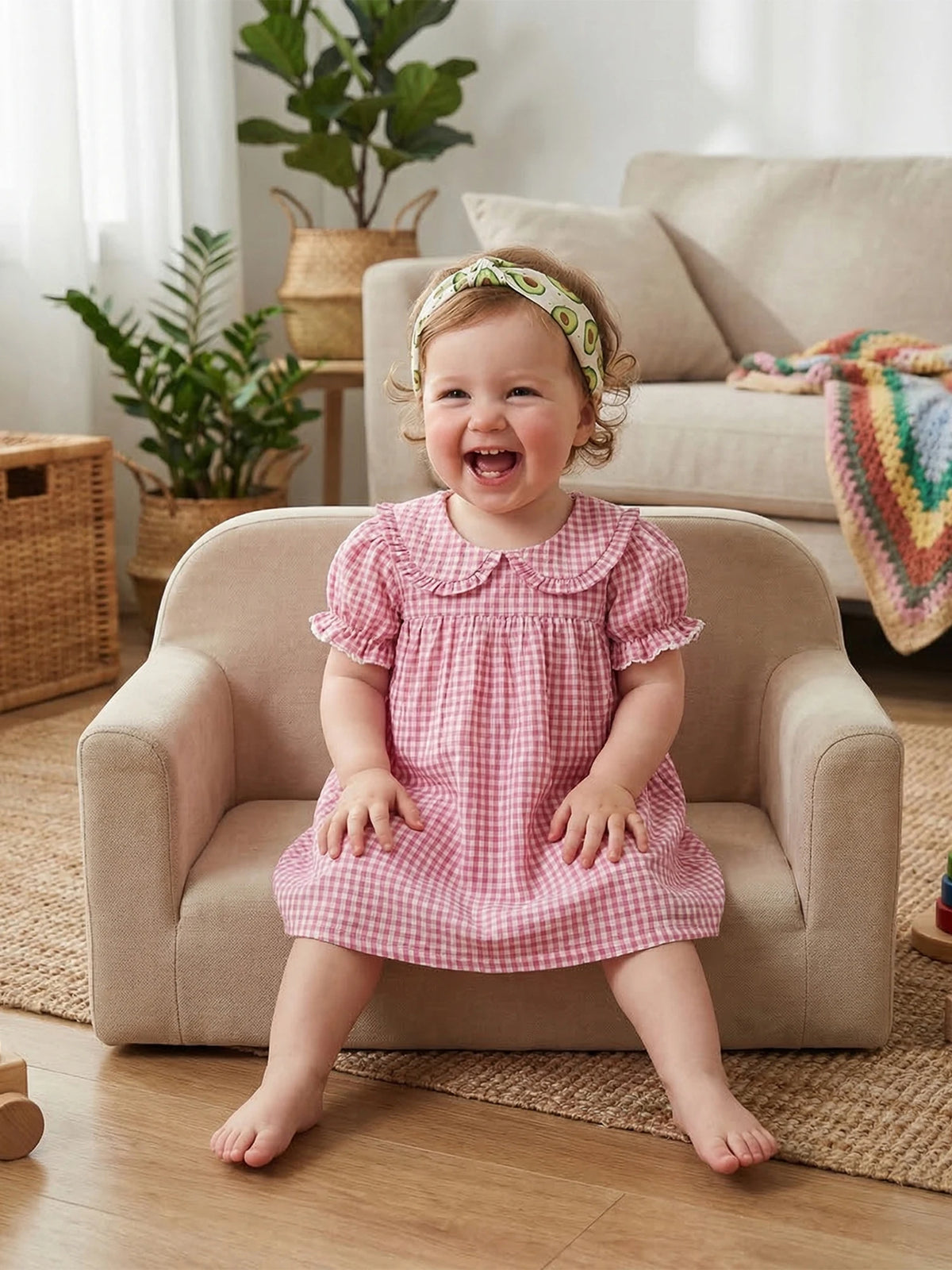 A toddler girl wearing a pink gingham A-line dress with a Peter Pan collar sitting on a sofa.