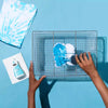 A hand is using a brush to apply blue liquid fabric dye onto white fabric inside a glass grid frame.