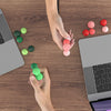 A hand is holding and arranging various colored soft sensory silicone massage balls on a wooden desk next to a laptop.