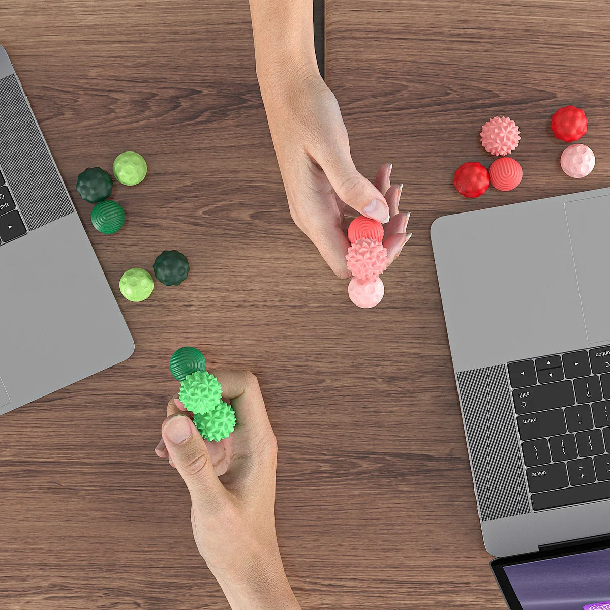 A hand is holding and arranging various colored soft sensory silicone massage balls on a wooden desk next to a laptop.