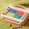 A pink magnetic drawing board for kids with colored magnetic shapes and a purple pencil lying nearby on green grass.