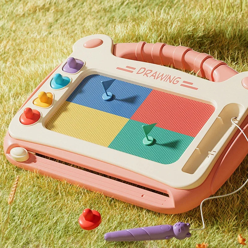 A pink magnetic drawing board for kids with colored magnetic shapes and a purple pencil lying nearby on green grass.