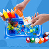 Hands are pouring colored liquid into a blue plastic tray filled with various gel molds and craft materials.
