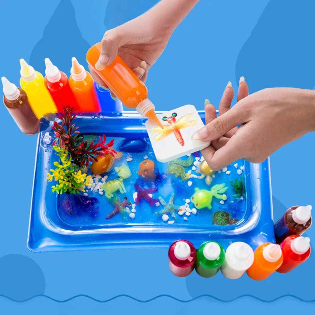 Hands are pouring colored liquid into a blue plastic tray filled with various gel molds and craft materials.