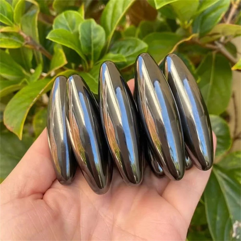 A hand holds several elongated, glossy black magnetic olive snake egg fidget toys against a green leafy background.