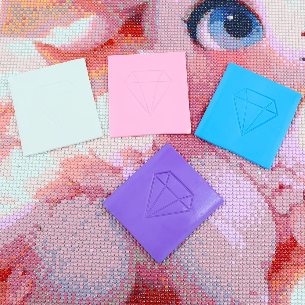 Four square blocks of diamond painting clay in pastel colors pink blue purple and white are arranged on a textured background