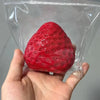 A hand holds a bright red strawberry squishy fidget toy sealed in clear plastic packaging.