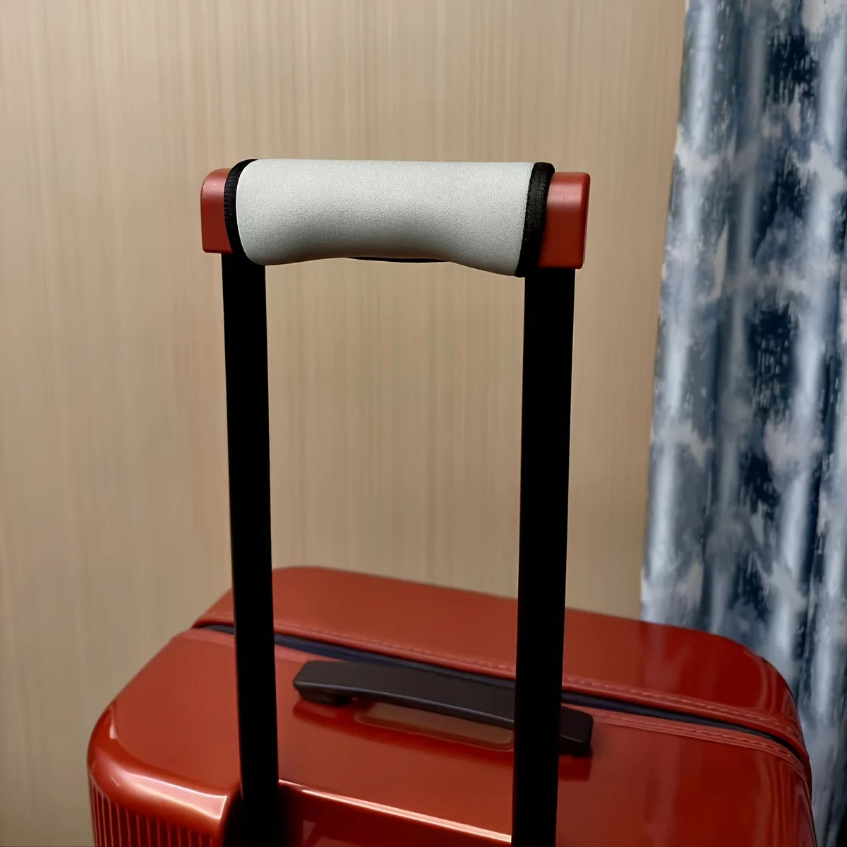 A close-up of a black luggage handle with a gray cushioned grip and a red suitcase visible in the background.
