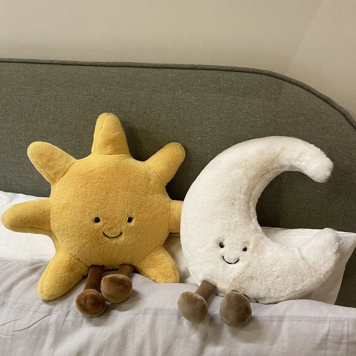 A yellow sun and white crescent moon plushie cushion set resting on a white bed with a gray headboard.