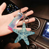 A hand holds a clear blue starfish beaded keychain charm against a dark car interior background.