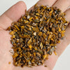 A hand holds a pile of natural tumbled stone chips in various shades of brown and gold.
