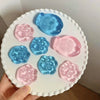 A hand holds a white plate with six pink and blue squishy cat paw toys.