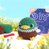A handmade green and brown bird amigurumi crochet kit featuring a small crocheted bird sitting on a white surface.