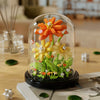 A glass display featuring an orange and yellow buildable eternal rose flower kit with green foliage.
