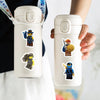 A hand holds a white cylindrical product with a lid and several cartoon ninja stickers attached.