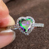 A close-up of a heart-shaped cubic zirconia ring sparkling on a finger against a dark brown background.