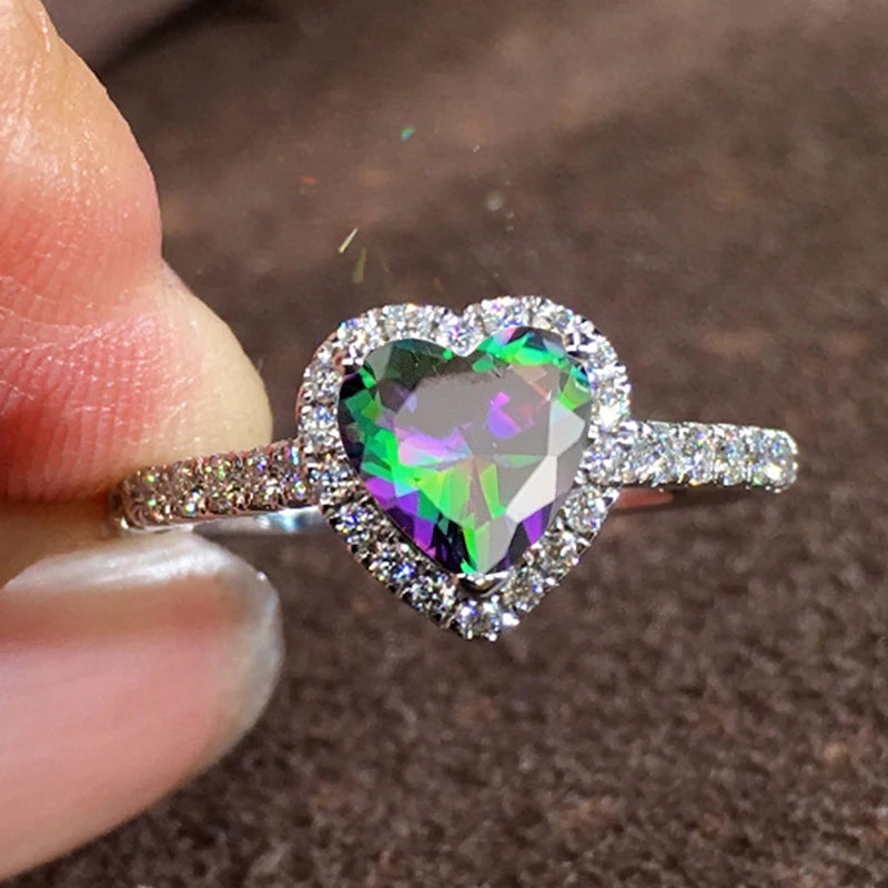 A close-up of a heart-shaped cubic zirconia ring sparkling on a finger against a dark brown background.