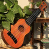 A wooden ukulele toy with adjustable tuning pegs rests against a green plant on a wooden shelf.