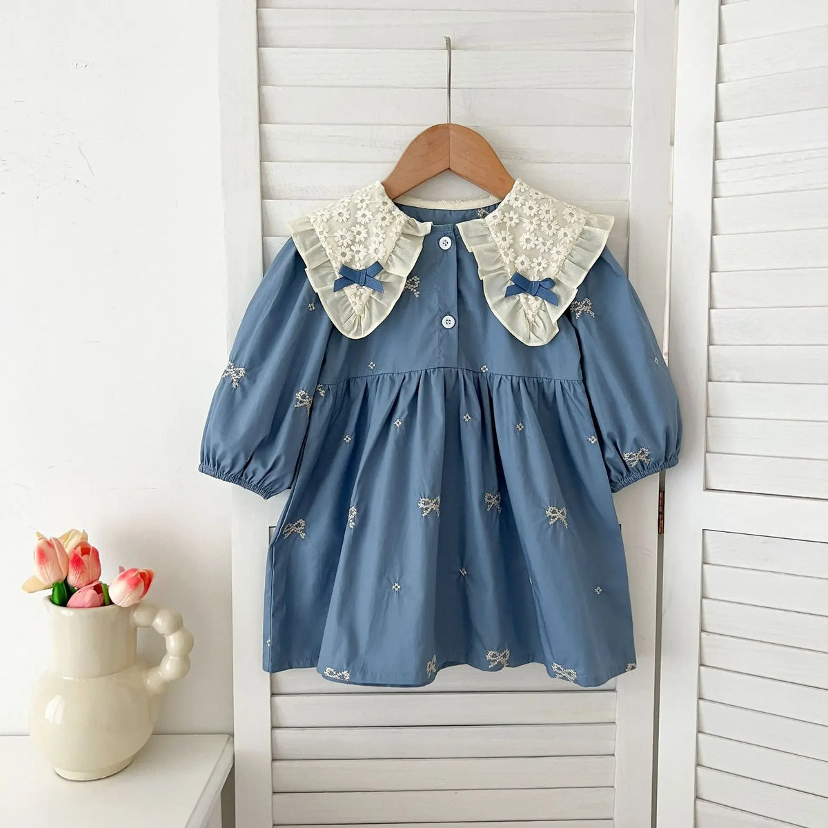 A blue cotton party dress for girls featuring lace collar details and embroidered accents, hanging on a wooden hanger.