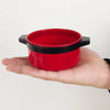 A bright red pretend play kitchen food set with a lid and black accents is held in a hand.
