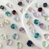 A collection of irregular square geometric glass beads in various shades of purple, blue, and green on a white surface.