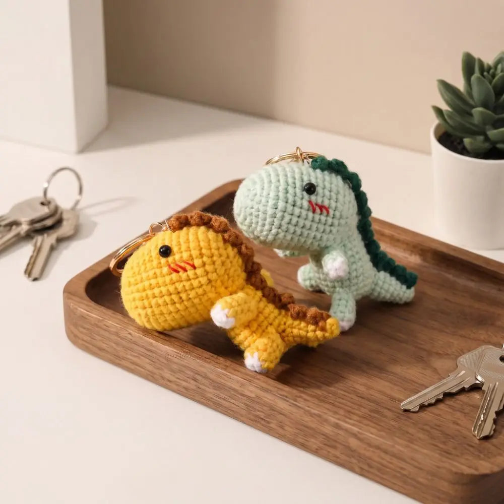 A yellow and green handmade crochet dinosaur keychain resting on a wooden tray with keys.