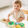 A smiling toddler holds a wooden busy board featuring musical and sensory activities.