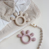 A light pink silicone baby teether with round beads rests on a beige cloth and white fabric.